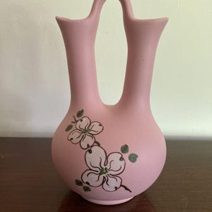 May include: A pink ceramic double vase with a handle, featuring a floral design of white flowers with green leaves.