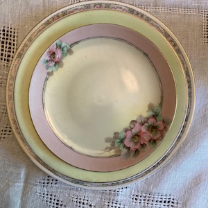 Puede incluir: Un juego de tres platos de porcelana vintage con un delicado diseño floral. Los platos presentan una gama de colores amarillo pálido, rosa y blanco con flores pintadas a mano.