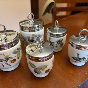 May include: A set of five white ceramic egg coddlers with shiny chrome lids featuring ring handles. The porcelain bodies are decorated with colorful botanical illustrations of strawberries, blackberries, and small birds perched on pine branches.
