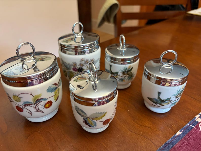 May include: A set of five white ceramic egg coddlers with shiny chrome lids featuring ring handles. The porcelain bodies are decorated with colorful botanical illustrations of strawberries, blackberries, and small birds perched on pine branches.