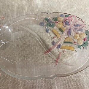 May include: Oval glass serving dish with a frosted finish and scalloped edges. The dish is decorated with a painted design featuring bells, a bow, and holly. The colors include pink, purple, yellow, and green.