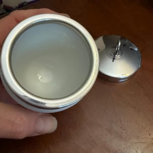 May include: A white ceramic container with a smooth interior and a metallic silver rim. Beside the container sits a matching circular metallic silver lid featuring a small, upright ring handle centered on its top surface for easy removal.