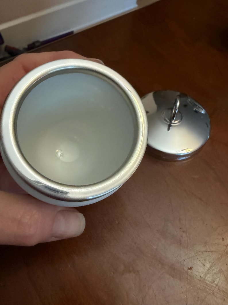 May include: A white ceramic container with a smooth interior and a metallic silver rim. Beside the container sits a matching circular metallic silver lid featuring a small, upright ring handle centered on its top surface for easy removal.