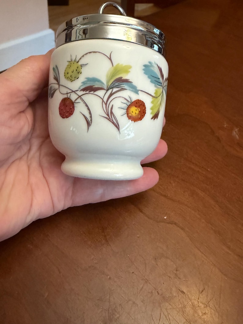 May include: A white porcelain egg coddler with a silver-toned metal lid and a ring handle. The body features a botanical pattern of red and yellow strawberries with green and blue leaves on thin brown stems, set against a smooth white surface.