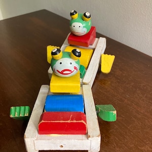 Pode incluir: Um comboio de brinquedo de madeira vintage com duas figuras de sapo. O comboio é feito de madeira branca com blocos coloridos. Os sapos têm corpos verdes, olhos pretos e bocas vermelhas. O comboio tem uma pega verde e branca.