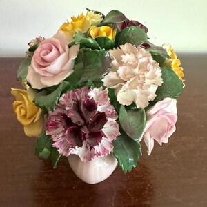 May include: A ceramic floral arrangement with roses, carnations, and other flowers in pink, yellow, and burgundy. The flowers are arranged in a small, pale pink vase. Green leaves provide a backdrop for the colourful blooms.