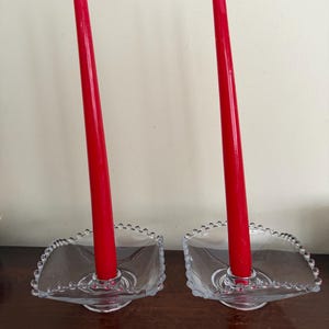 Könnte beinhalten: Zwei klare Glas-Kerzenhalter mit roten Kerzen. Die Kerzenhalter haben eine quadratische Form mit einem Perlenrand. Die Kerzen sind hoch und schlank, mit einem weißen Docht oben. Die Kerzenhalter stehen auf einer dunkelbraunen Oberfläche.