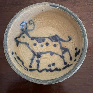 May include: A round, shallow ceramic bowl with a tan, textured surface. The interior features a hand-painted, dark blue line drawing of a goat with spots, curved horns, and a small beard, surrounded by simple decorative blue markings along the rim.