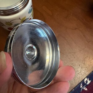 May include: A circular metal lid with a polished silver finish and a central recessed knob, shown from the underside. In the background, a white ceramic container features a colorful botanical pattern of strawberries and green leaves.