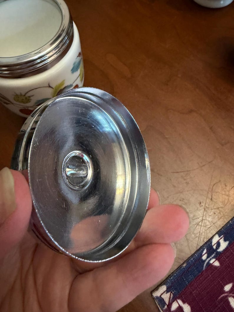 May include: A circular metal lid with a polished silver finish and a central recessed knob, shown from the underside. In the background, a white ceramic container features a colorful botanical pattern of strawberries and green leaves.