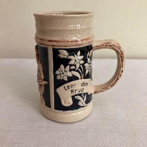 Pode incluir: Um caneco de cerveja de cerâmica com borda e alça bege, e corpo azul escuro. O caneco apresenta desenhos florais brancos, uma faixa com o texto "Leer den Krug" e um relevo de uma pessoa. A alça é marrom.