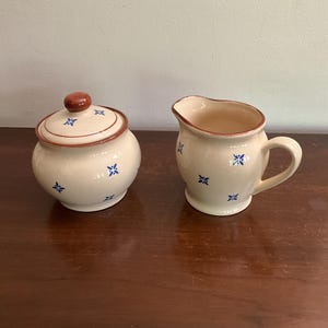 May include: A cream-colored ceramic sugar bowl with a lid and a matching creamer. Both pieces have a brown rim and a blue and white floral pattern.