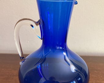 May include: A cobalt blue glass pitcher with a clear glass handle. The pitcher has a rounded body and a narrow neck.