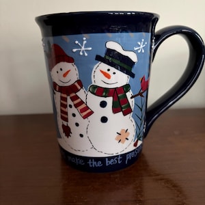 May include: A ceramic mug with a navy blue rim and handle. The mug features a light blue background with two snowmen wearing hats and scarves. The text "to make the best present" is written on the bottom of the mug.