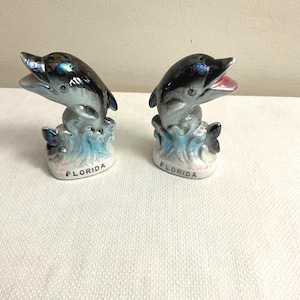 May include: A pair of ceramic dolphin salt and pepper shakers. The dolphins are jumping out of the water and have iridescent blue and black paint. The base of each shaker says "Florida".