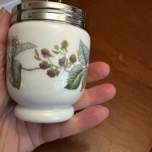 May include: A white ceramic egg coddler with a shiny silver metal lid and a small ring handle. The body of the coddler features a botanical illustration of green leaves and clusters of purple and green blackberries on a thin brown vine.
