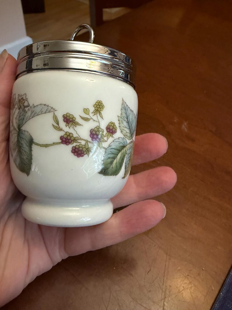 May include: A white ceramic egg coddler with a shiny silver metal lid and a small ring handle. The body of the coddler features a botanical illustration of green leaves and clusters of purple and green blackberries on a thin brown vine.