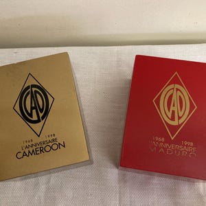 May include: Two rectangular boxes, one gold and one red, with a diamond-shaped logo and text. The gold box reads "1968 L'ANNIVERSAIRE 1998 CAMEROON." The red box reads "1968 L'ANNIVERSAIRE 1998 MADURO."