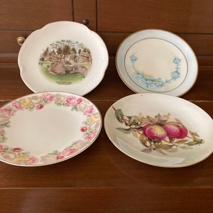 May include: Four decorative plates with different designs. One plate features a scenic image of a park with the text "Scenic Dell Lodge, Salisbury, Pa.", another plate has a floral design with pink and yellow roses, a third plate has a blue floral design, and the last plate has a painting of peaches on a branch.