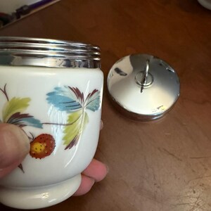 May include: A white ceramic jar with a silver-toned metal rim and a matching circular lid featuring a small handle. The jar is decorated with a painted pattern of green and blue leaves alongside a single orange strawberry with yellow seeds.