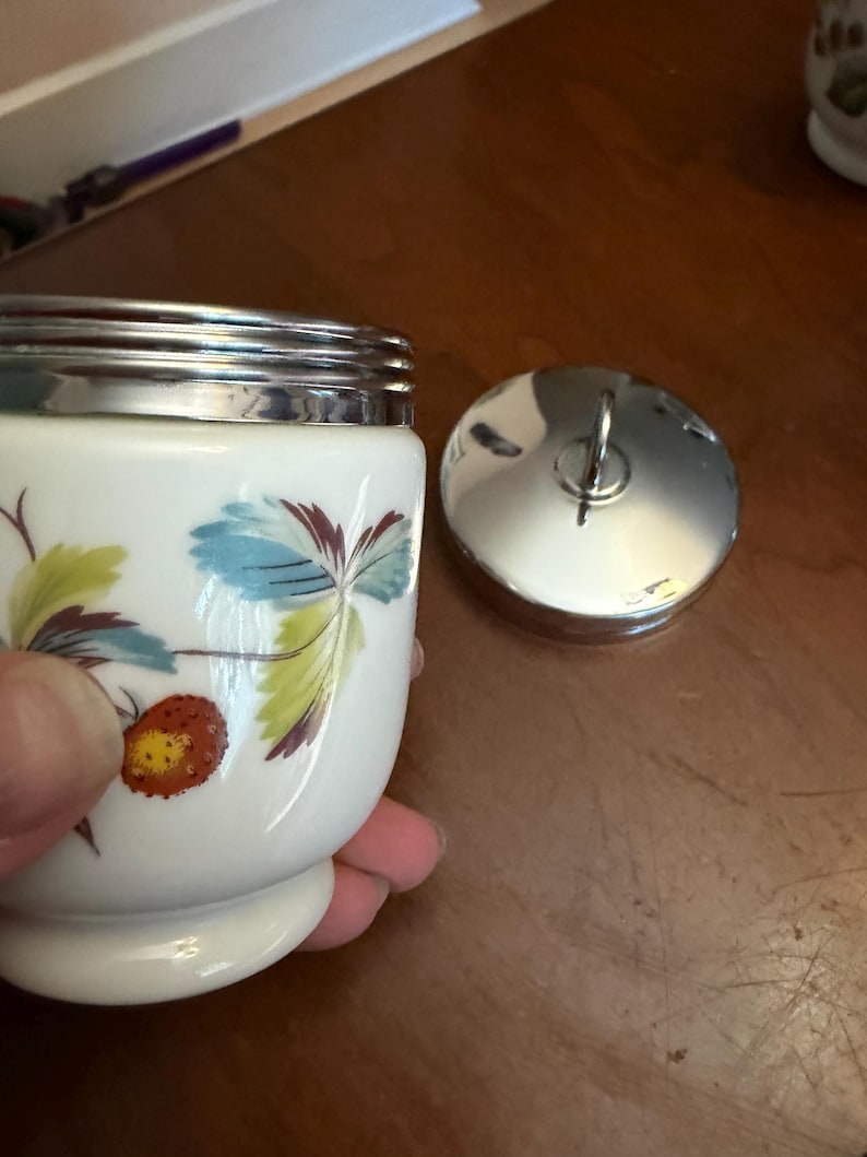 May include: A white ceramic jar with a silver-toned metal rim and a matching circular lid featuring a small handle. The jar is decorated with a painted pattern of green and blue leaves alongside a single orange strawberry with yellow seeds.
