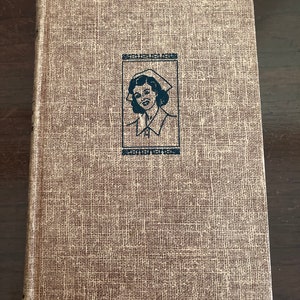 May include: A vintage book with a brown linen cover featuring a black and white illustration of a nurse wearing a white cap and uniform.