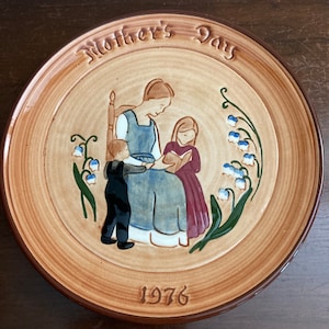May include: A brown ceramic plate featuring a raised design of a mother reading to two children. The plate is inscribed with the words "Mother's Day" and "1976".