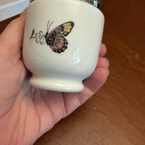 May include: A white porcelain egg coddler with a silver-toned metal lid and a ring handle. The body features a colorful butterfly graphic with yellow, black, and red wing details, positioned next to a small stylized blue and brown floral design.