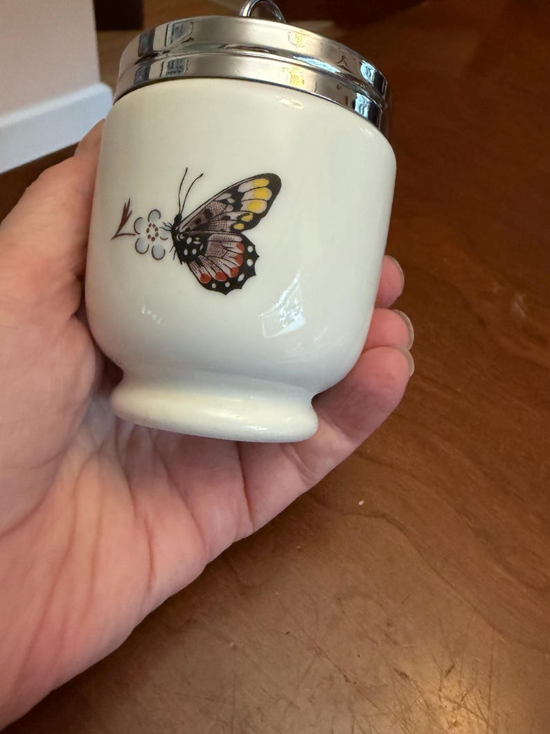 May include: A white porcelain egg coddler with a silver-toned metal lid and a ring handle. The body features a colorful butterfly graphic with yellow, black, and red wing details, positioned next to a small stylized blue and brown floral design.