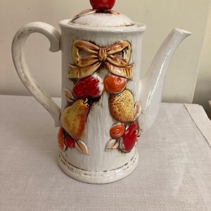 May include: White ceramic teapot with a fruit and bow design. The teapot features a red apple on the lid, a bow, and various fruits in relief. The handle is curved, and the spout is long and slender.