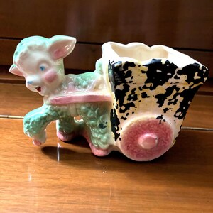 Könnte beinhalten: Ein Keramik-Pflanztopf in Form eines Lamms, das einen Wagen zieht. Das Lamm ist grün, rosa und weiß mit schwarzen Augen. Der Wagen ist schwarz, weiß und gelb mit einem rosafarbenen Rad.