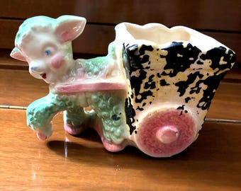May include: A ceramic planter shaped like a lamb pulling a wagon. The lamb is green, pink, and white with black eyes. The wagon is black, white, and yellow with a pink wheel.