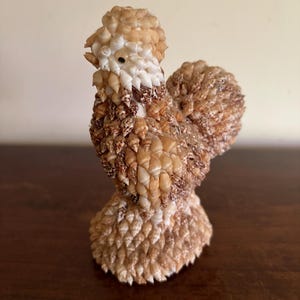 May include: A decorative figurine shaped like a chicken, constructed entirely from numerous small, overlapping seashells in shades of tan, beige, and white. The sculpture features a rounded body, a textured tail, and a single dark bead for an eye.