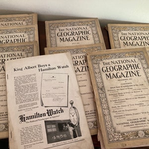 May include: A stack of vintage National Geographic magazines from the early 1900s. The covers feature illustrations and text, including "King Albert Buys a Hamilton Watch" and "The Geographic's Notable Americans".