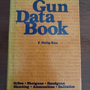 May include: A hardback book titled Outdoor Life Gun Data Book by F. Philip Rice. The cover is a tan and brown colour with dark blue text. The front features a list of technical topics including rifles, shotguns, handguns, and ballistics.