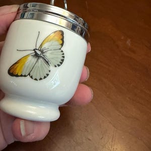May include: A white porcelain egg coddler featuring a detailed illustration of a butterfly with yellow and black wing tips. The container has a polished silver-tone metal lid with a circular handle loop at the top and a flared base design.