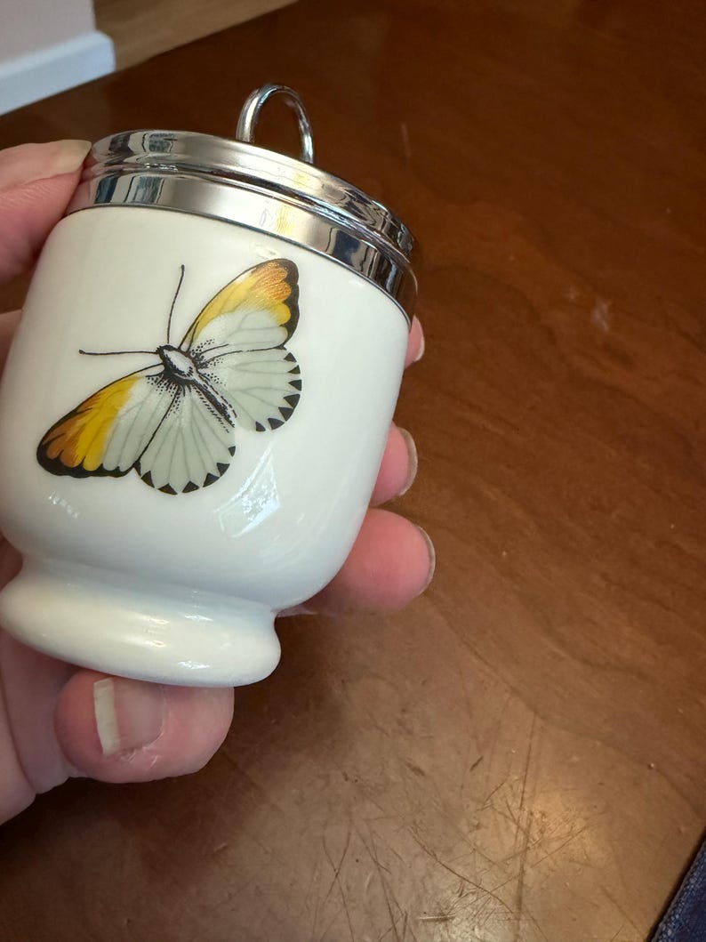May include: A white porcelain egg coddler featuring a detailed illustration of a butterfly with yellow and black wing tips. The container has a polished silver-tone metal lid with a circular handle loop at the top and a flared base design.