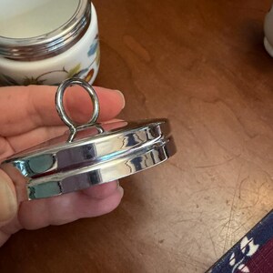 May include: A circular silver-toned metal lid featuring a central ring-shaped handle. The lid has a stepped, tiered edge design with a polished, reflective finish. It is designed to fit onto a matching white ceramic jar with a silver rim.