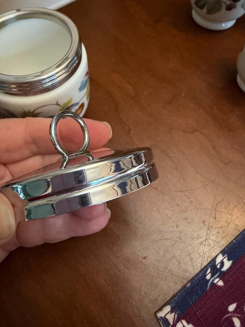 May include: A circular silver-toned metal lid featuring a central ring-shaped handle. The lid has a stepped, tiered edge design with a polished, reflective finish. It is designed to fit onto a matching white ceramic jar with a silver rim.