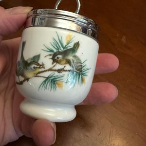 May include: A white porcelain egg coddler with a polished silver-tone metal lid and a circular handle. The body features a painted illustration of two small birds perched on green pine branches with yellow accents against a plain white background.