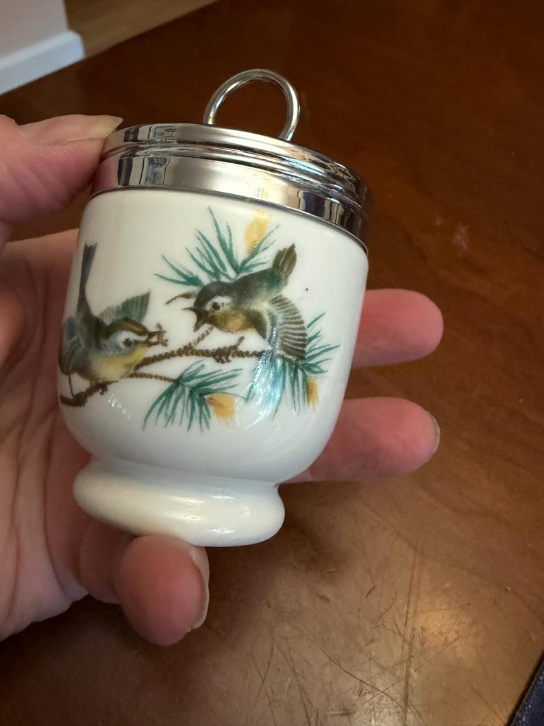 May include: A white porcelain egg coddler with a polished silver-tone metal lid and a circular handle. The body features a painted illustration of two small birds perched on green pine branches with yellow accents against a plain white background.