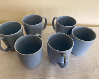 May include: Six light blue ceramic mugs with rounded handles. The mugs are arranged in a semi-circle on a light gray surface.