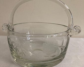 Könnte beinhalten: Klarer Glaskorb mit gebogenem Griff und geätzten floralen Designs. Der Korb hat eine runde Schalenform mit zwei kleinen dekorativen Griffen auf beiden Seiten. Die geätzten Designs umfassen Blumen und Blätter.