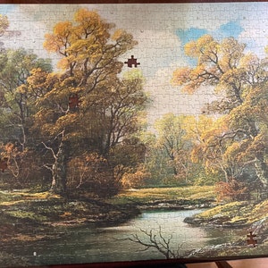 May include: A completed jigsaw puzzle showing an autumnal landscape with a river, trees, and colourful foliage. Several puzzle pieces are missing, revealing the wooden backing. The puzzle is set in a wooden frame.