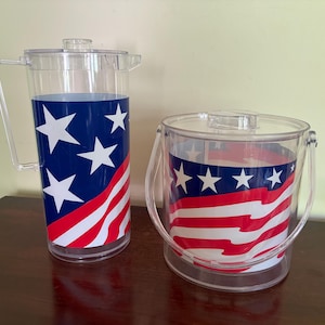May include: A clear acrylic pitcher with a handle and lid alongside a matching clear acrylic ice bucket with a swing handle. Both items feature a wrap-around graphic design of a blue field with white stars and red and white horizontal stripes.