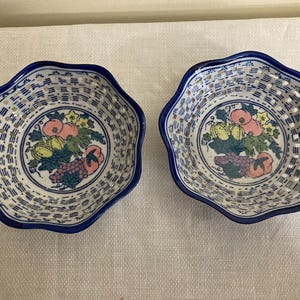 May include: Two decorative ceramic bowls with a blue scalloped edge and a white and blue lattice design. Each bowl features a central fruit illustration with pink, yellow, and purple elements. These bowls are ideal for serving snacks or displaying small items.