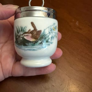 May include: A white porcelain egg coddler with a silver-toned metal lid featuring a circular handle. The body of the coddler is decorated with a detailed illustration of a small brown bird perched near green reeds and blue water.