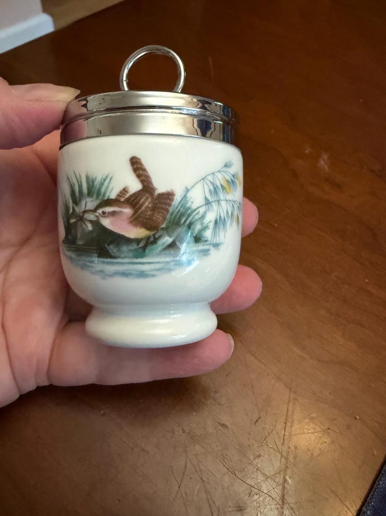 May include: A white porcelain egg coddler with a silver-toned metal lid featuring a circular handle. The body of the coddler is decorated with a detailed illustration of a small brown bird perched near green reeds and blue water.