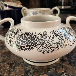 May include: White ceramic creamer with a black floral design. The creamer has a round body and two handles.