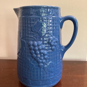 May include: A blue ceramic pitcher with a grapevine design. The pitcher has a textured surface and a handle.